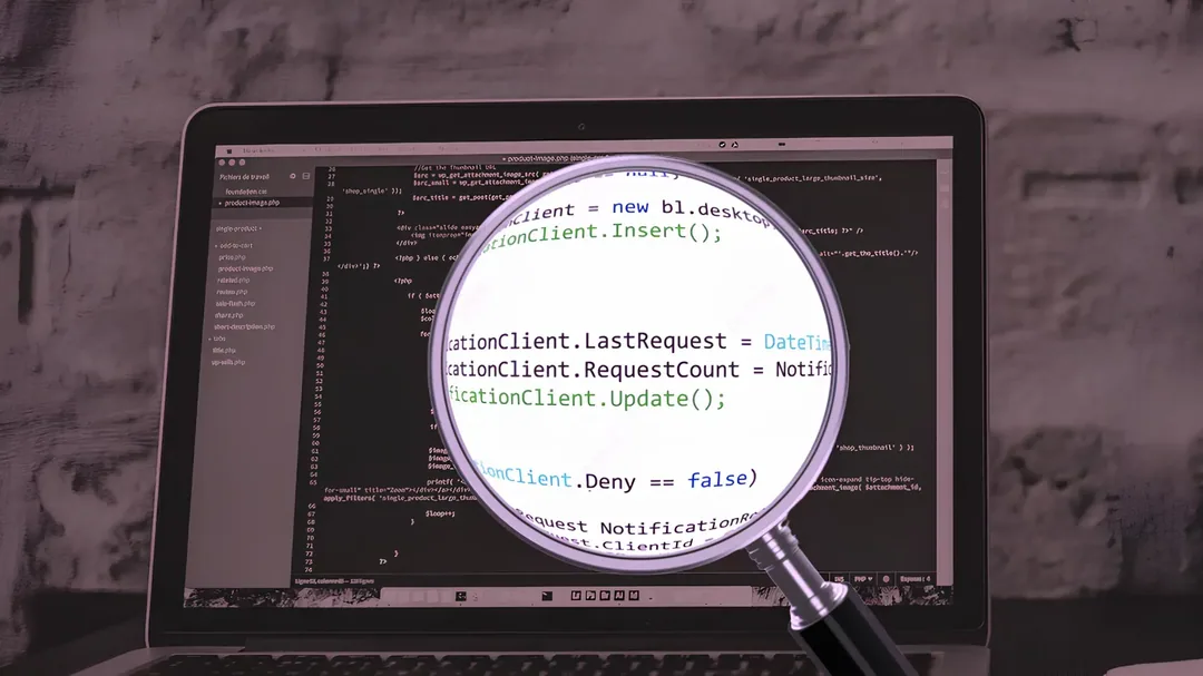 Black-and-white image of a laptop displaying code, with a magnifying glass enlarging text. Conveys focus on software development and coding details.